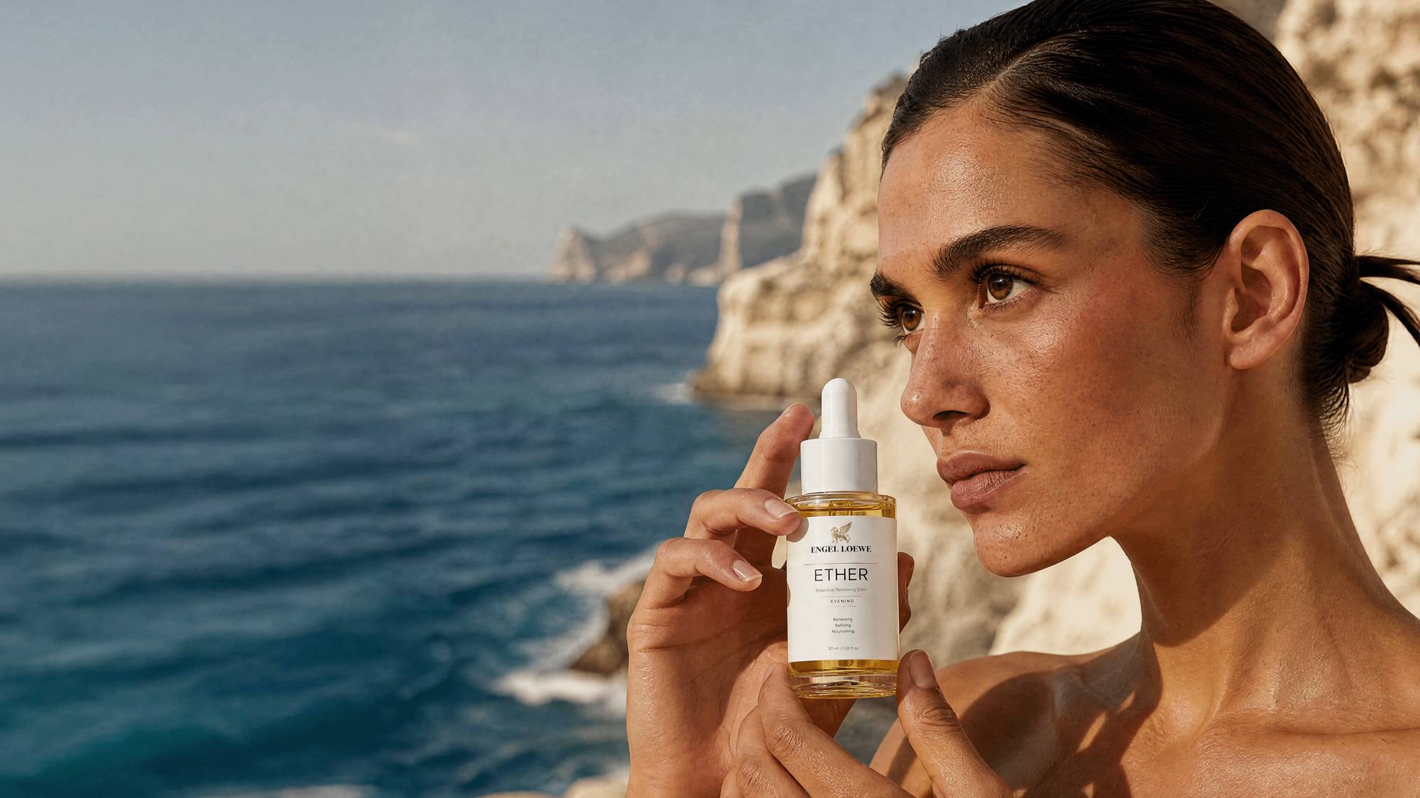 ETHER facial oil held against Mediterranean cliffs